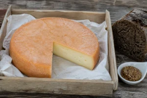 Whole orange-rinded abbey cheese disc with one wedge cut away in a market crate - Abbaye du Mont des Cats | KnowTheCheese