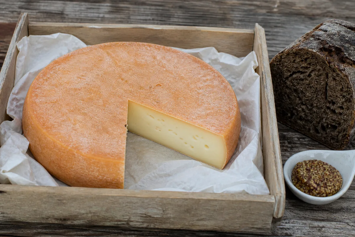 Whole orange-rinded abbey cheese disc with one wedge cut away in a market crate - Abbaye du Mont des Cats | KnowTheCheese