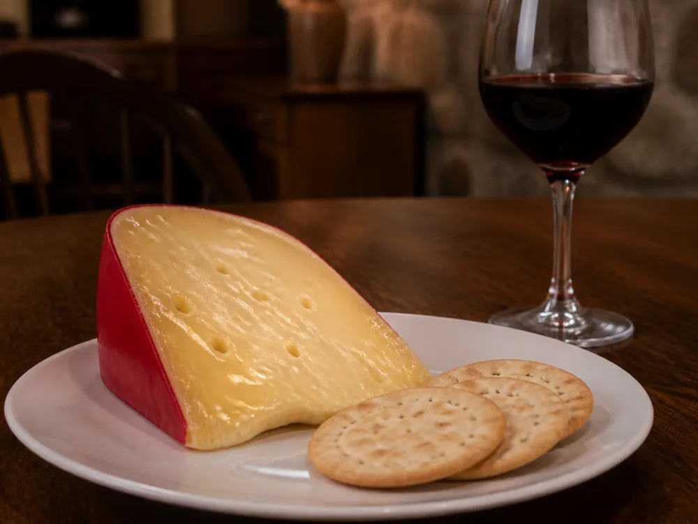 Wedge with its red wax rim on a plain plate with water crackers and a glass of red wine - Airedale | KnowTheCheese