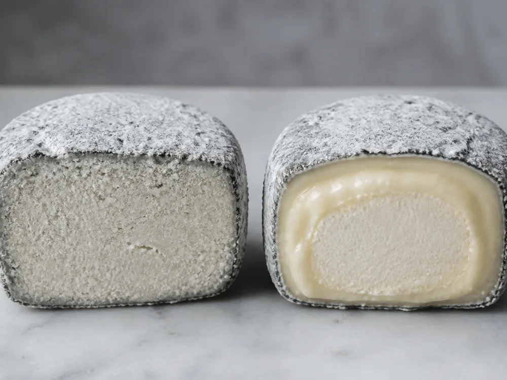 Two ash-covered cheeses cut open, one firm and pale throughout and one with a soft creamy band - Aisy Cendré | KnowTheCheese