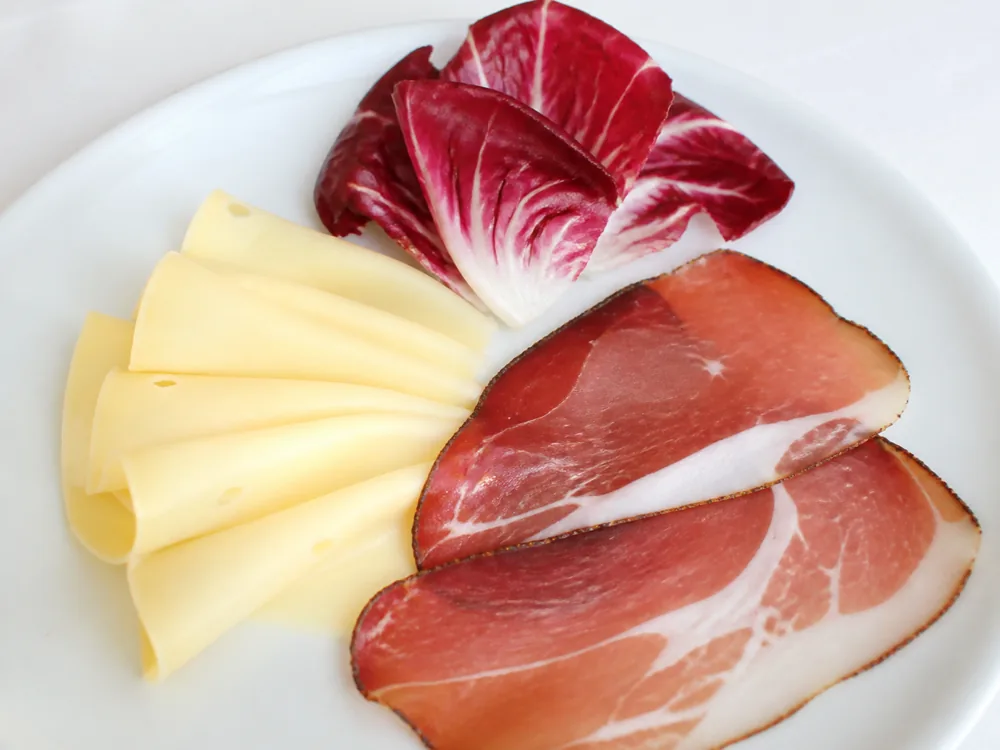 Thin slices of straw-gold cheese fanned on a plate beside speck and radicchio leaves - Alta Badia | KnowTheCheese