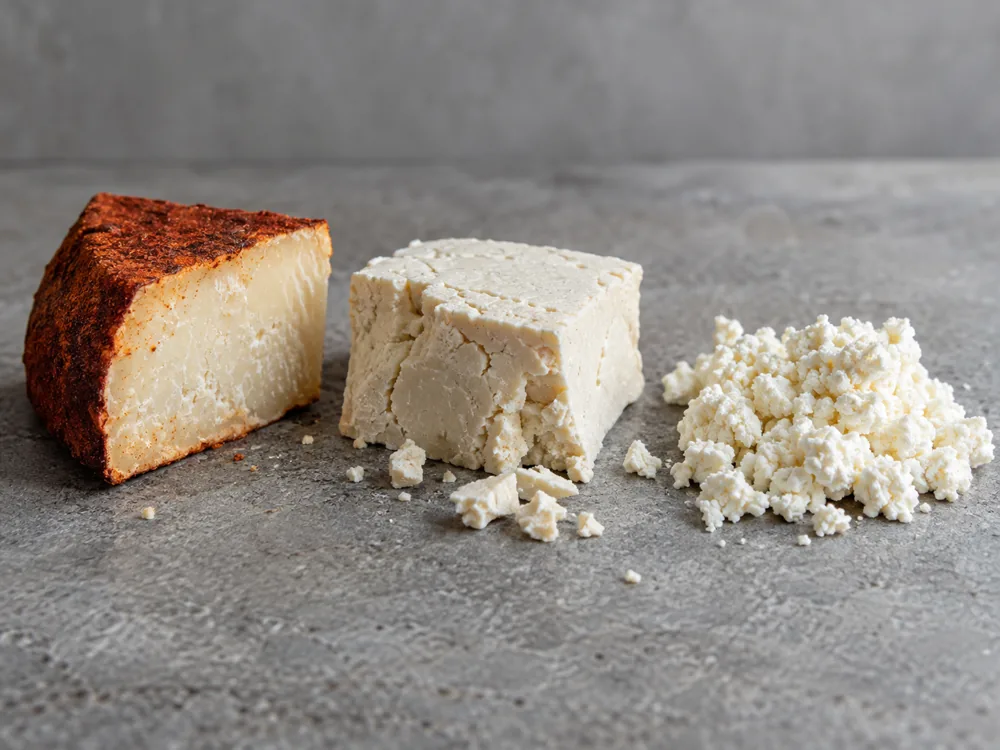 Red-coated aged cheese wedge beside a dry white block and fresh white crumbles - Añejo Enchilado | KnowTheCheese