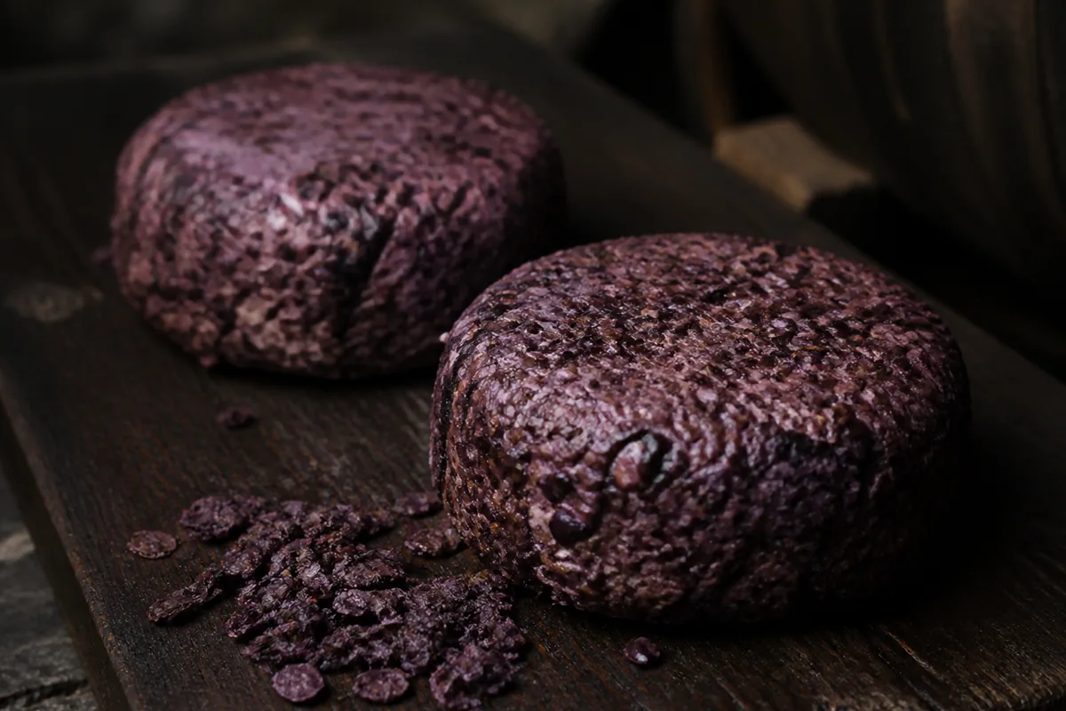 Two small flat discs encrusted with purple grape skins and pips on a dark board - Aromes au Gene de Marc | KnowTheCheese