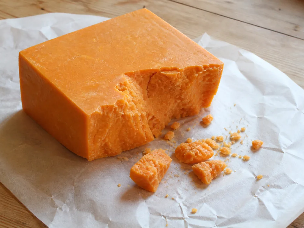 Broken chunk of deep orange cheese on waxed paper on a wooden kitchen table with crumbs beside it - Auld Lochnagar | KnowTheCheese
