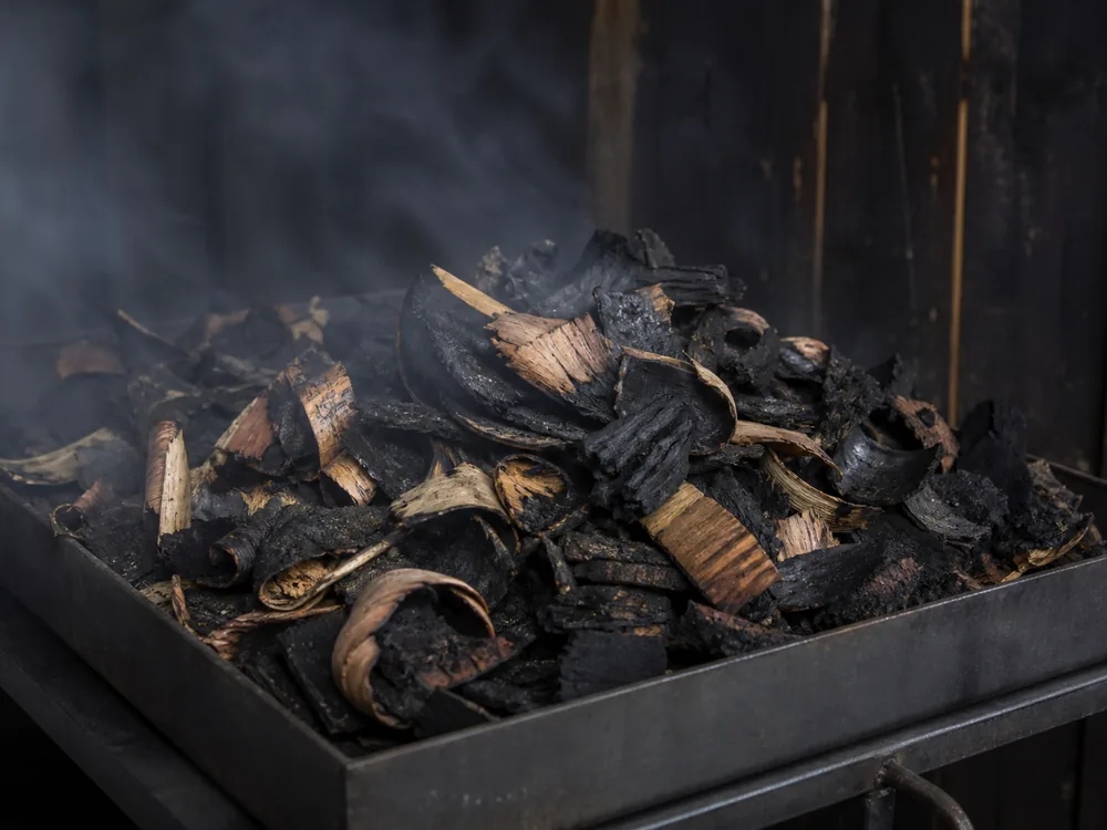 Coarse charred oak barrel shavings on a metal tray in a cold smokehouse with thin smoke drifting - Auld Reekie | KnowTheCheese