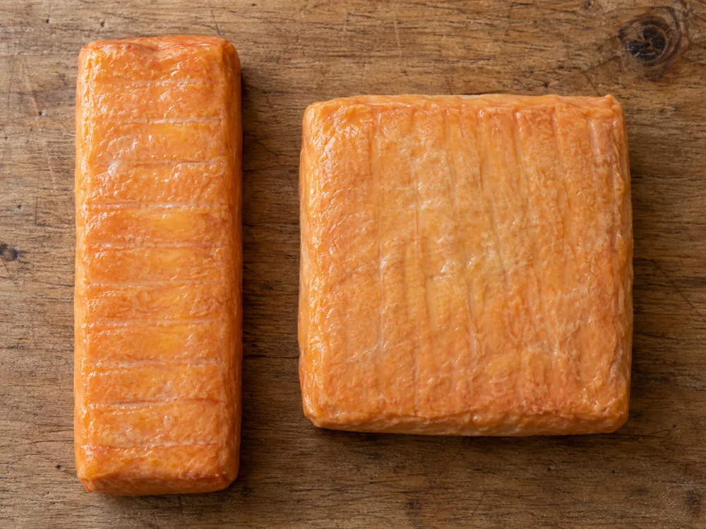 Top view of a long narrow washed-rind bar beside a flat square washed-rind block, both with an orange rind - Baguette laonnaise | KnowTheCheese