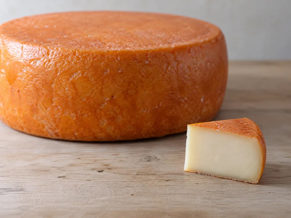 Small cut wedge with ivory paste in front of a whole orange-rinded wheel - Baluchon | KnowTheCheese