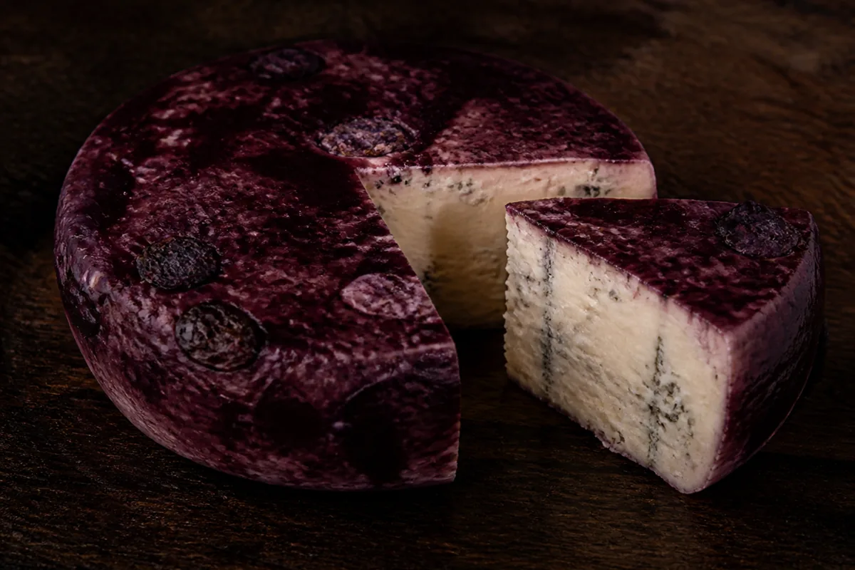 Squat wheel with a deep purple wine-stained surface and a wedge showing ivory paste with fine blue veins - Basajo | KnowTheCheese