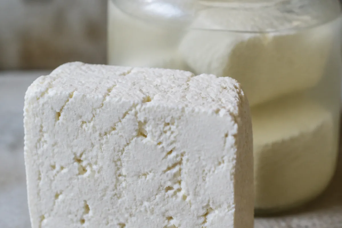 Rough-cut block of chalk-white cheese full of small holes standing beside a plain jar of cloudy brine - Batzos | KnowTheCheese
