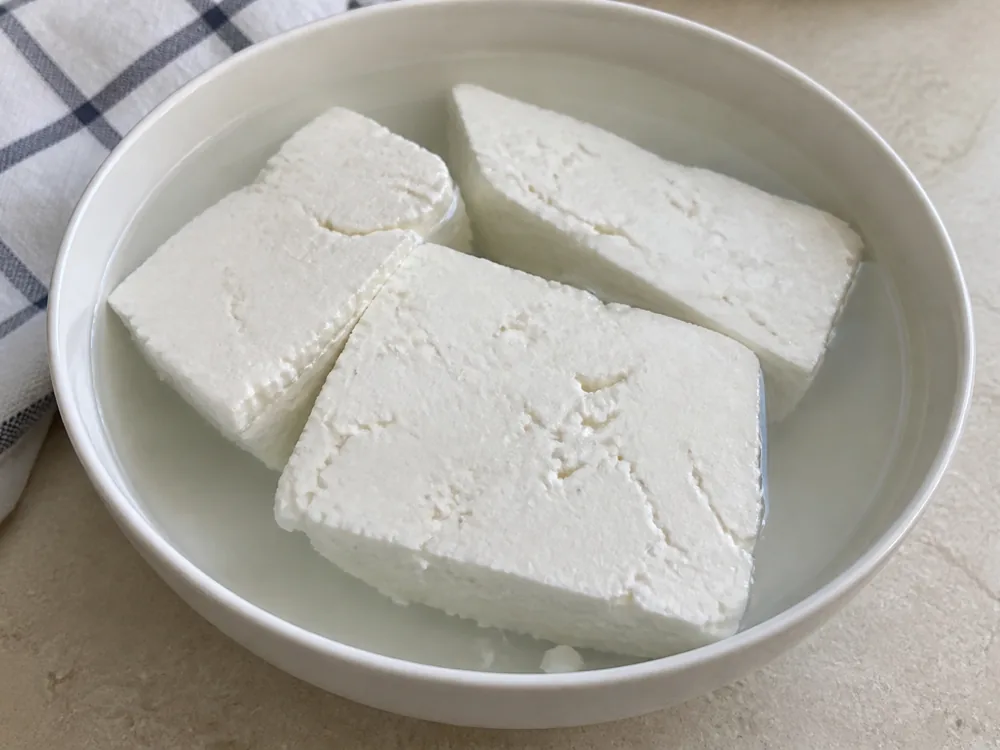 Bowl of cold water with several white cheese slabs submerged and a folded cloth beside it - Batzos | KnowTheCheese