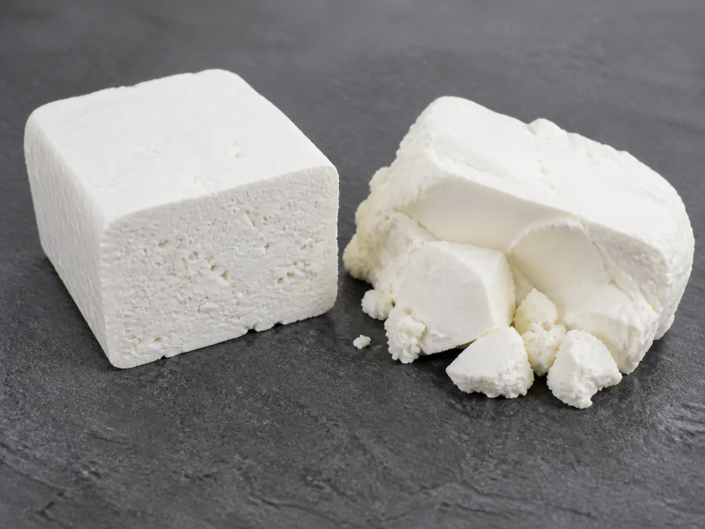 Firm white block holding a square cut face beside a softer white cheese broken into crumbs on grey slate - Batzos | KnowTheCheese