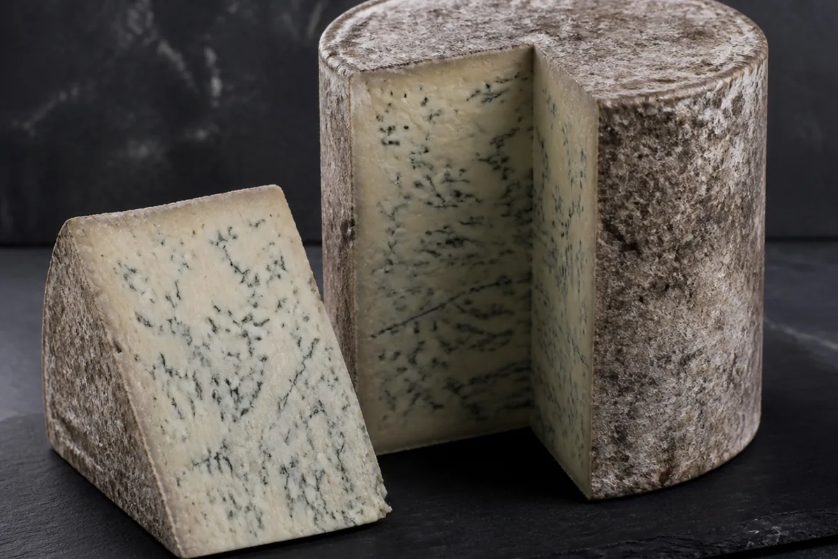 Tall cylindrical blue cheese with a dry grey-brown natural crust and a wedge showing fine even veining - Bayley Hazen Blue | KnowTheCheese