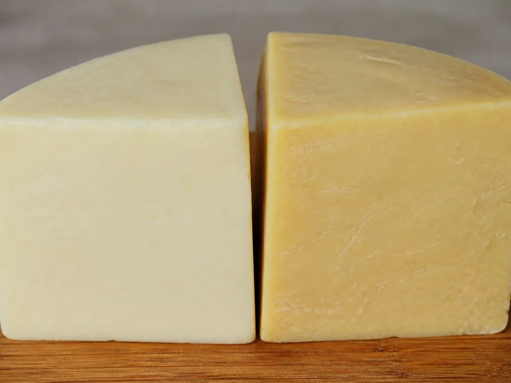Two cut faces of the same cheddar side by side, the left paler and smoother and the right deeper gold with fine fracture lines - Baylough | KnowTheCheese