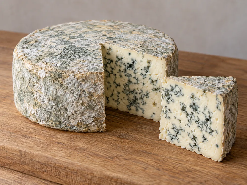Whole flat Bleu d'Auvergne cylinder with a blue-green tinged crust and a cut wedge showing ivory paste with blue-green veins - Bleu d'Auvergne | KnowTheCheese