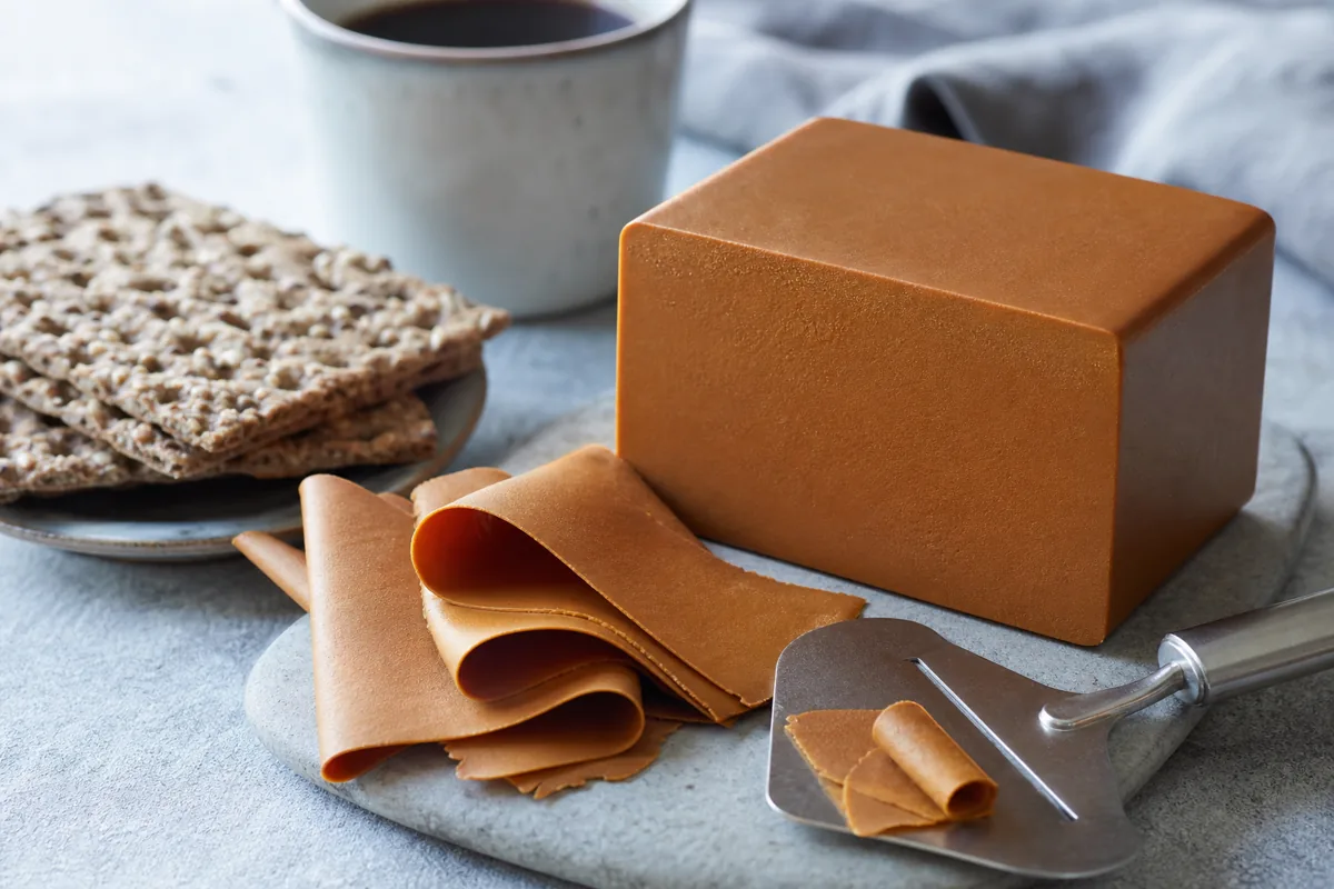 Brown Brunost block with thin curled slices and a cheese plane - Brunost | KnowTheCheese
