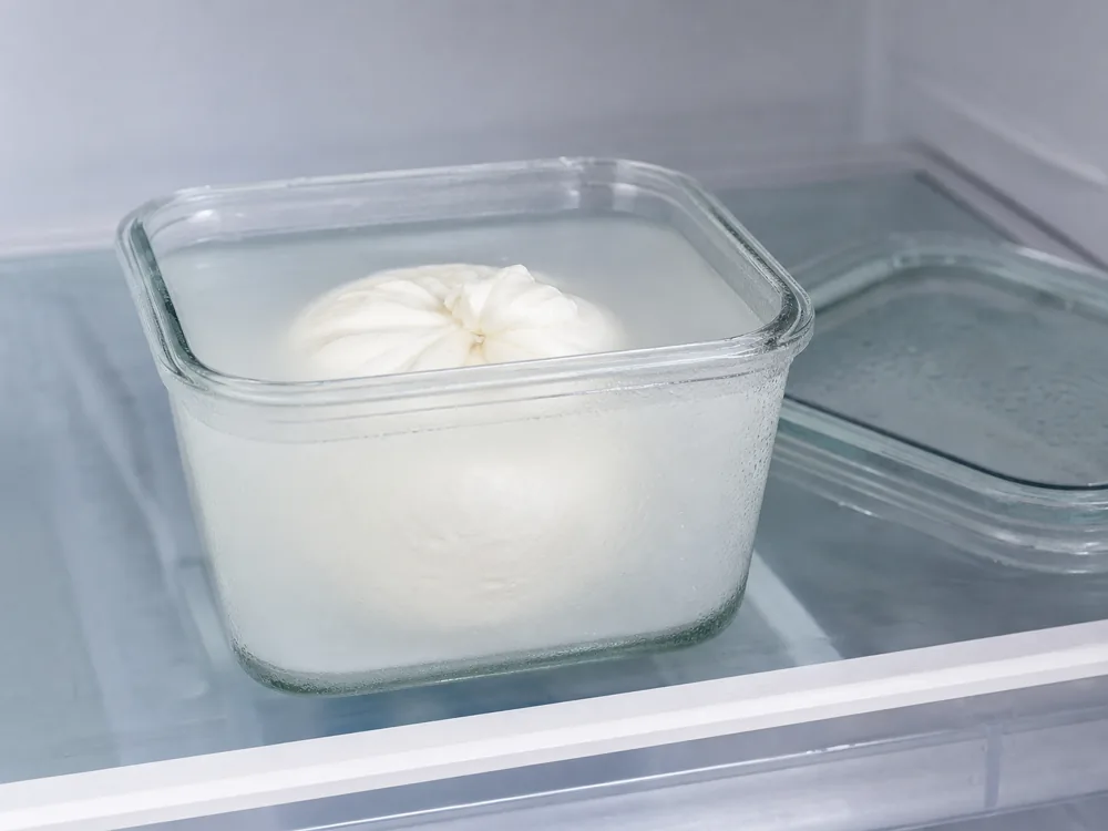 Whole burrata stored under its packing liquid in a glass container - Burrata | KnowTheCheese