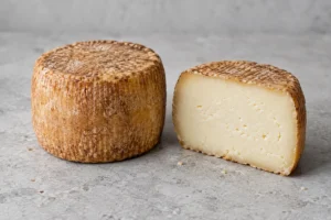 Whole small cylindrical cheese with a ridged malt-brown rind beside a halved one with pale paste - Caciobirraio | KnowTheCheese