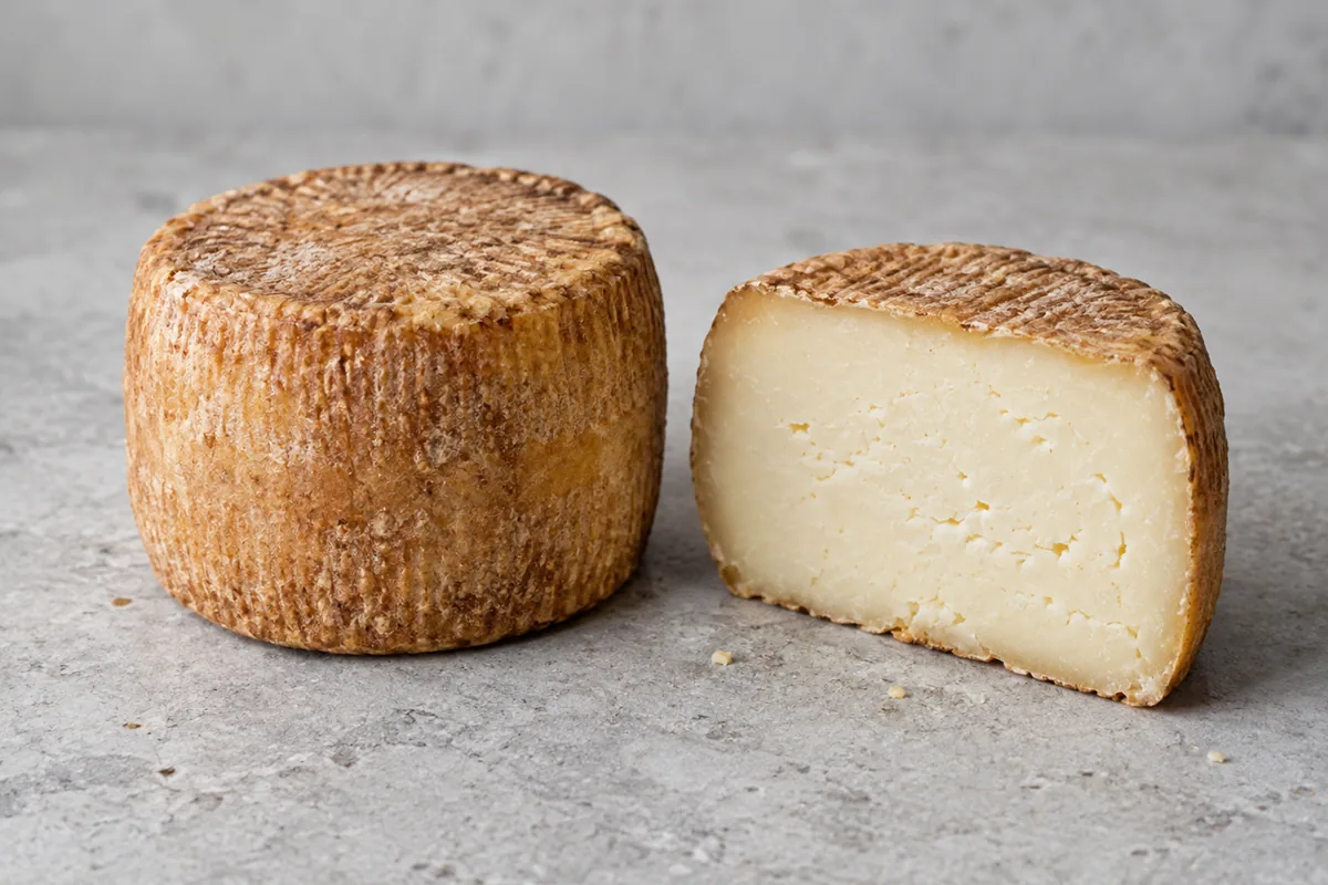 Whole small cylindrical cheese with a ridged malt-brown rind beside a halved one with pale paste - Caciobirraio | KnowTheCheese