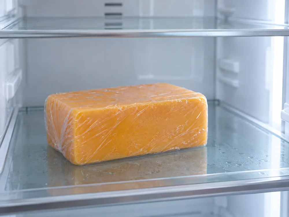 Orange Cheddar block wrapped closely in clear plastic film on a refrigerator shelf - Canadian Cheddar cheese | KnowTheCheese