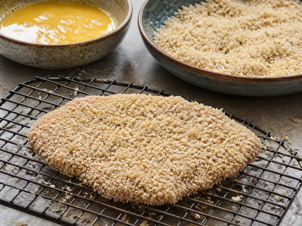 A raw cutlet coated in coarse dry breadcrumbs on a rack beside bowls of egg and crumbs - chicken parmesan | KnowTheCheese
