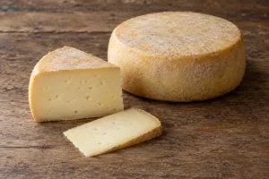 Whole round cheese and a cut wedge with a slice on a wooden table - Colony Cheese | KnowTheCheese
