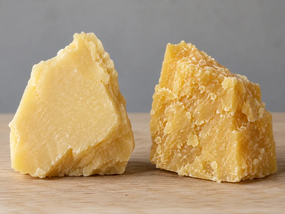 Two broken chunks of hard cheese on a wooden board, the left paler with a smooth surface and the right deeper yellow with a crumbly, flaky surface - Parmigiano-Reggiano vs Parmesan | KnowTheCheese