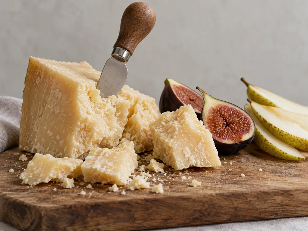 Wedge of hard granular cheese broken into chunks with a small knife, beside halved figs and pear slices on a wooden board - Parmigiano-Reggiano vs Parmesan | KnowTheCheese