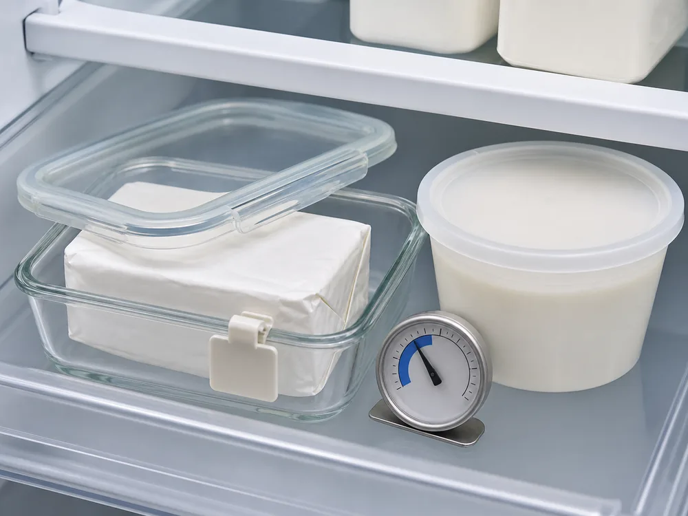 Wrapped cream cheese brick in a glass container beside a sealed tub and refrigerator thermometer - Cream Cheese | KnowTheCheese