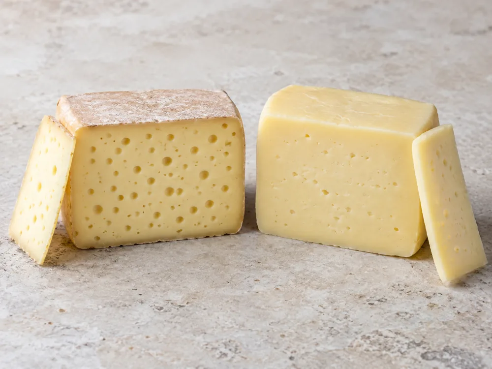 Two cut blocks of semi-soft cheese side by side, one with a thin rind and many small round eyes and one rindless with very small irregular openings - Danbo | KnowTheCheese