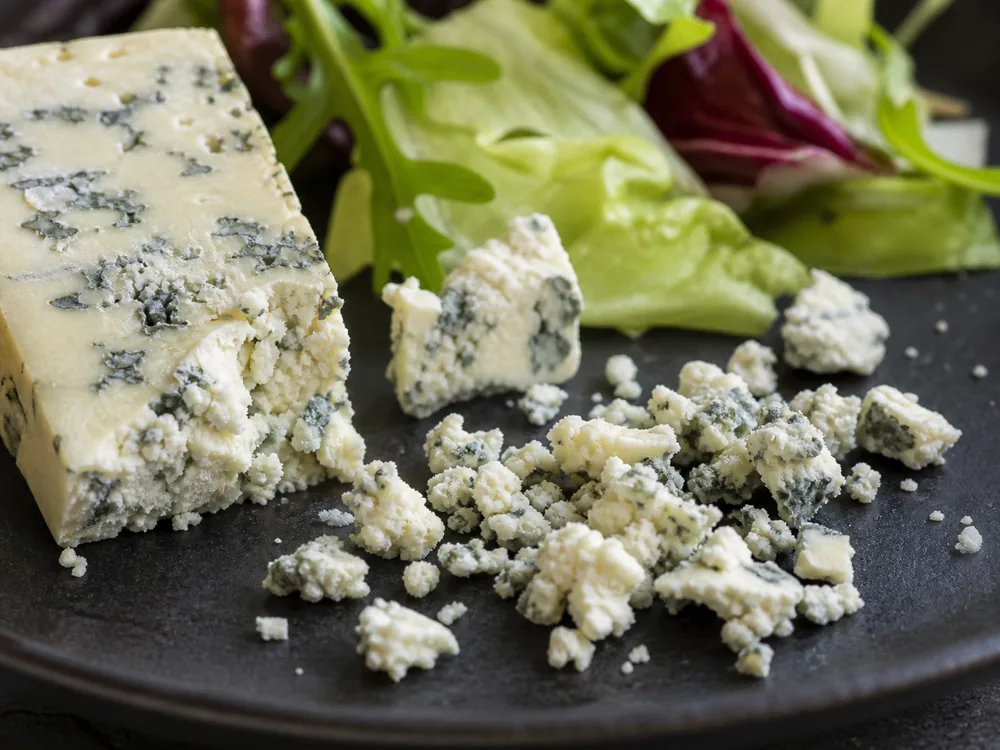 Uneven Danish Blue crumbles beside salad leaves and a blue-veined wedge - Danish Blue | KnowTheCheese