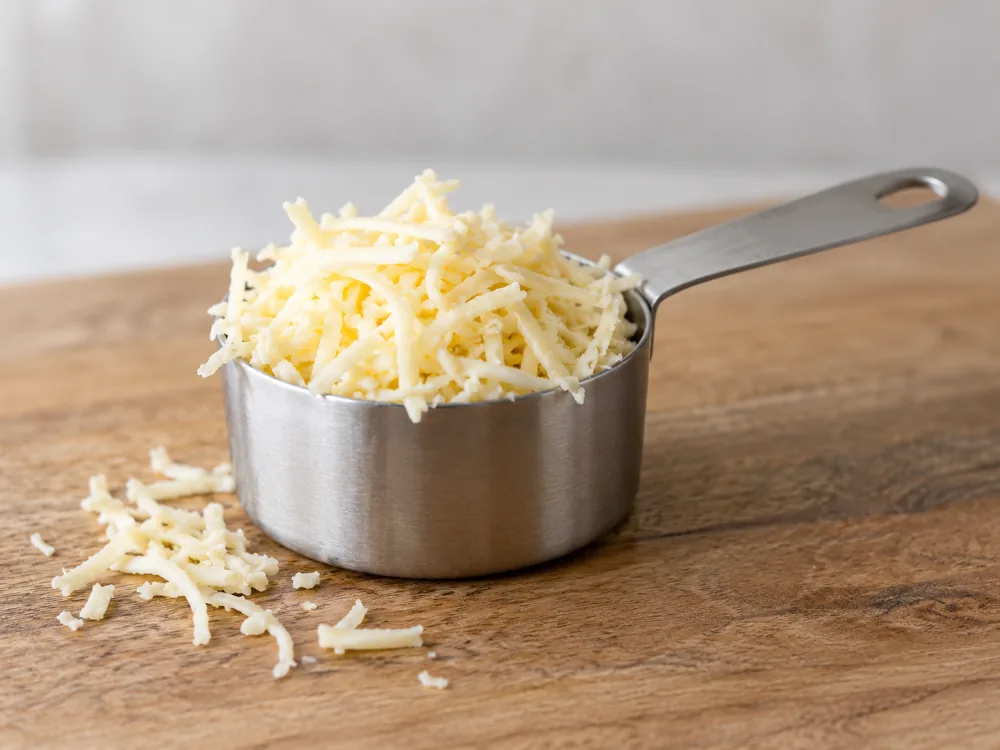Steel measuring cup of shredded pale yellow cheese on a wooden board - Dubliner cheese | KnowTheCheese