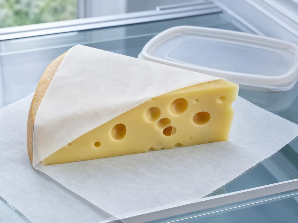 Cut Emmental-style wedge partly covered with breathable cheese paper on a refrigerator shelf - Emmental | KnowTheCheese