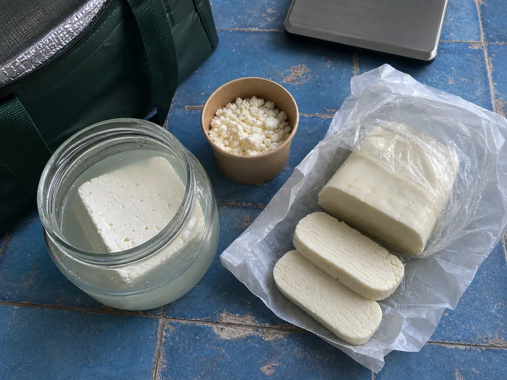 Feta block in brine, dry feta crumbles, and thick halloumi slices on a blue tile counter - Feta vs Halloumi | KnowTheCheese