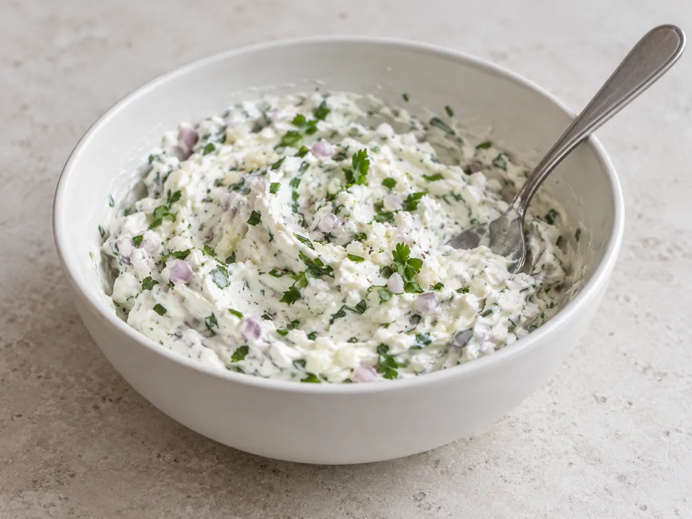 Thick white fromage blanc mixed with chopped shallots and green herbs in a white bowl with a spoon - Fromage Blanc | KnowTheCheese