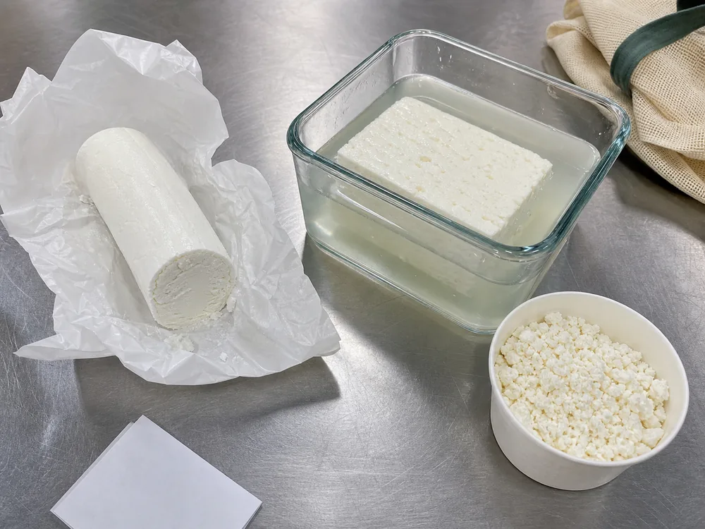 Fresh goat cheese log, feta block in brine, and dry pre-crumbled feta on a deli counter - Goat Cheese vs Feta | KnowTheCheese