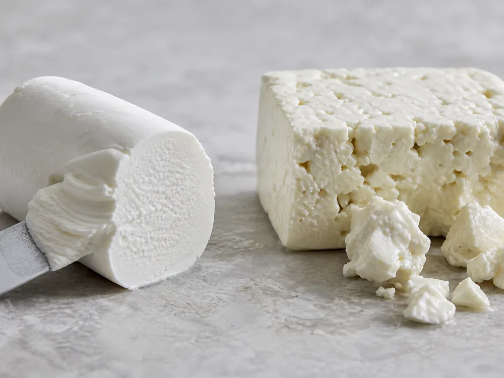 Rindless fresh goat cheese with a creamy cut face beside a fissured feta block and broken chunks - Goat Cheese vs Feta | KnowTheCheese