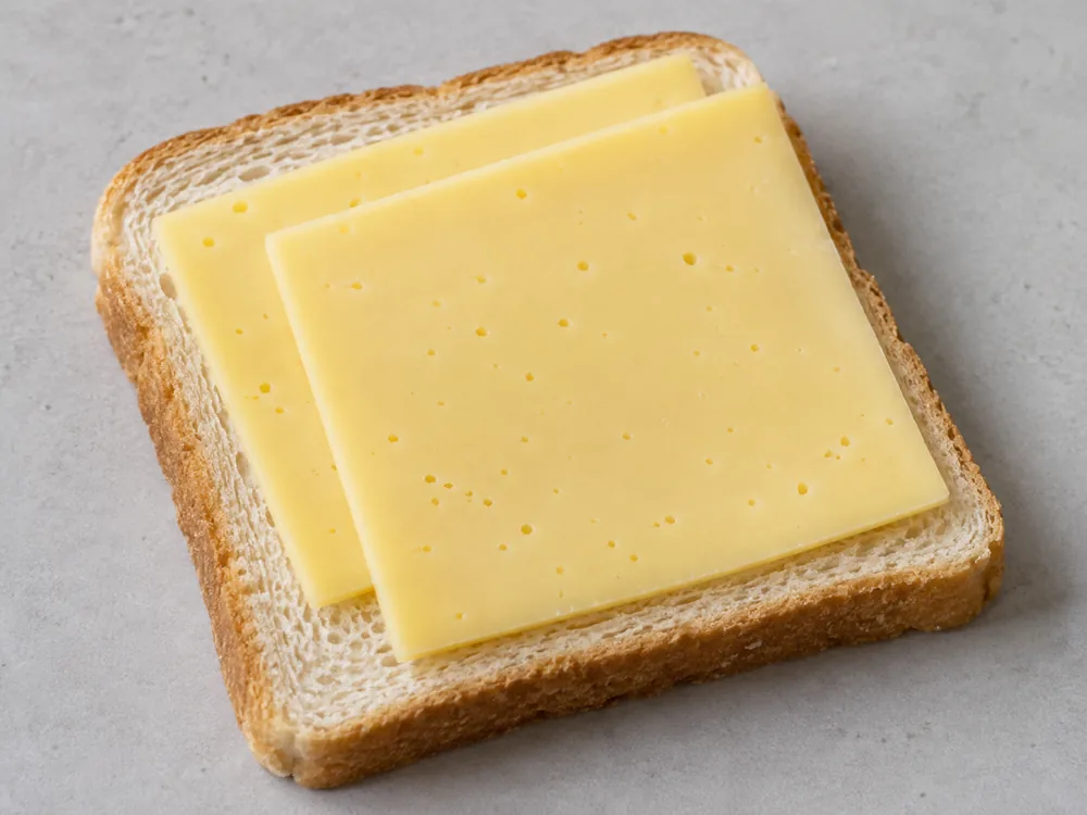 Two slices of pale yellow cheese with a few tiny holes on a slice of bread - Hushållsost | KnowTheCheese