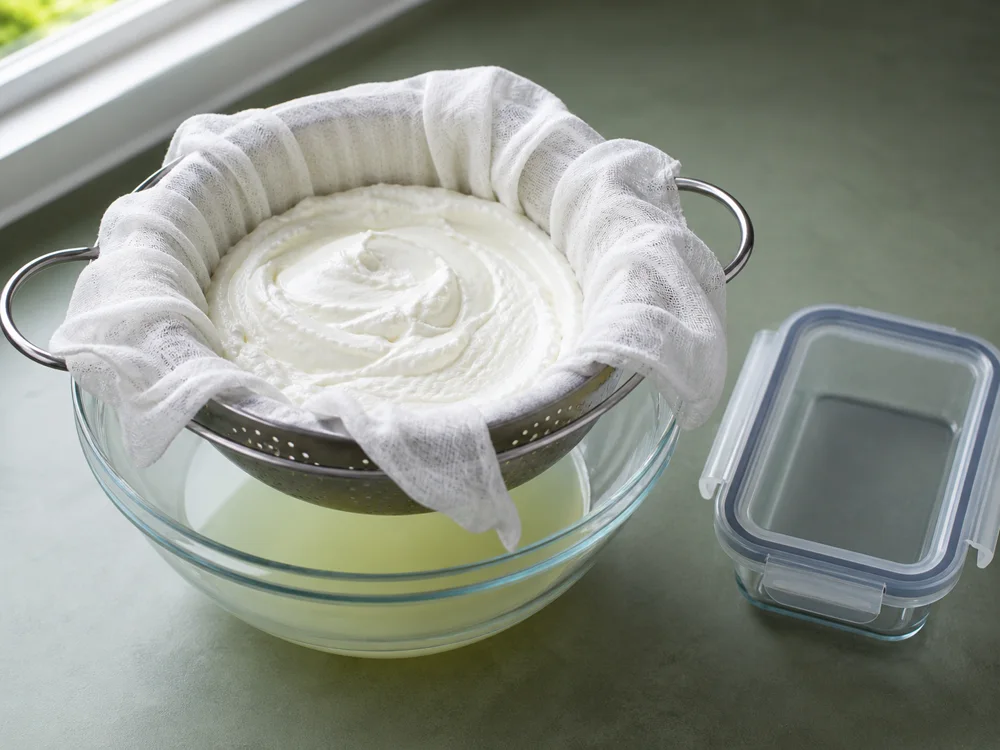 Cheesecloth-lined colander draining labneh above a bowl of whey - Labneh | KnowTheCheese