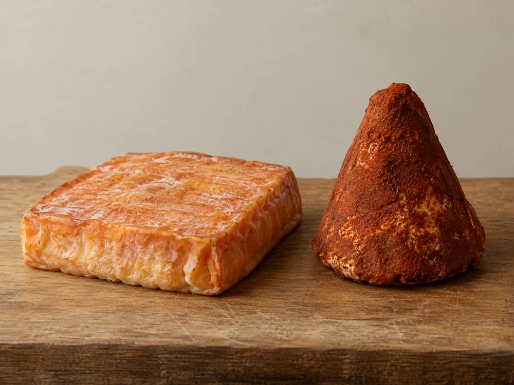 A flat square washed-rind cheese beside a cone-shaped cheese coated in red-brown paprika - Maroilles | KnowTheCheese
