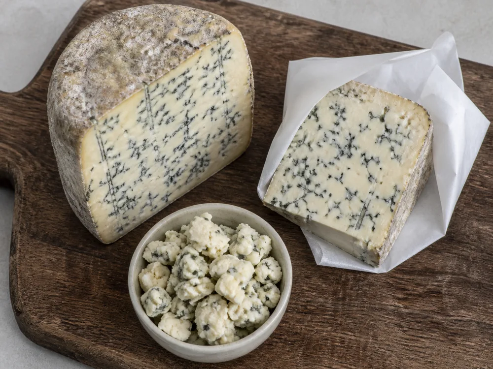 A wheel section, a paper-wrapped wedge, and a bowl of crumbles of the same blue cheese - Maytag Blue | KnowTheCheese