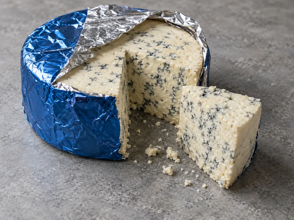 A round blue cheese wheel in decorative foil, partly unwrapped, with one wedge cut out - Maytag Blue | KnowTheCheese