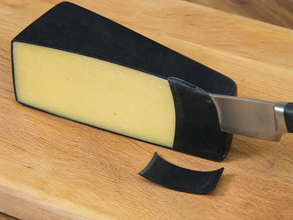 A knife cutting through black wax on a firm cheese wedge with a wax curl beside it - Midnight Moon | KnowTheCheese
