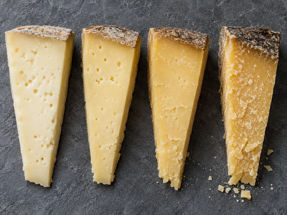 Four wedges of the same cooked-curd cheese in a row showing an ageing progression from pale to deep golden - Montasio | KnowTheCheese