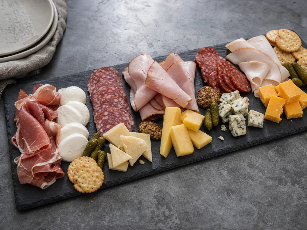 Meat and cheese board with prosciutto, salami, ham, cured sausage, turkey, mozzarella, provolone, gouda, blue cheese, cheddar, pickles, and crackers.