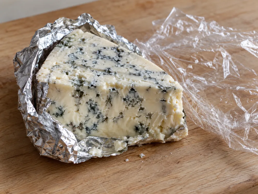 Wedge of blue cheese half wrapped in aluminium foil beside a sheet of cling film - Gorgonzola storage | KnowTheCheese