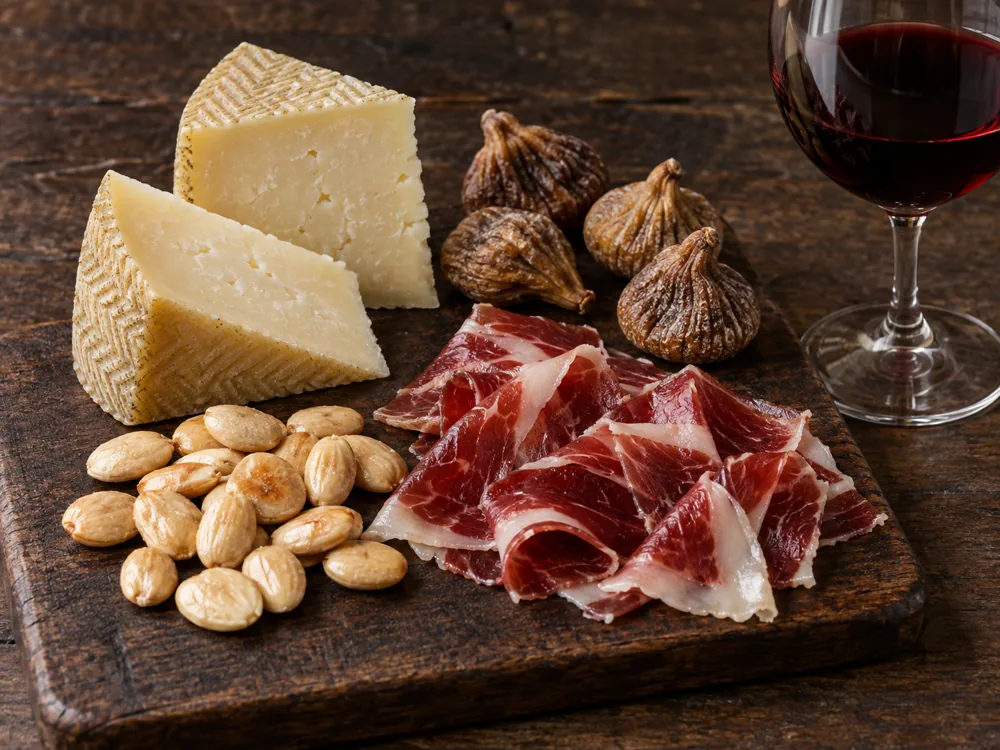 Two Manchego wedges with pale Marcona almonds, thin slices of cured ham, dried figs and a glass of red wine - Manchego Pairing | KnowTheCheese