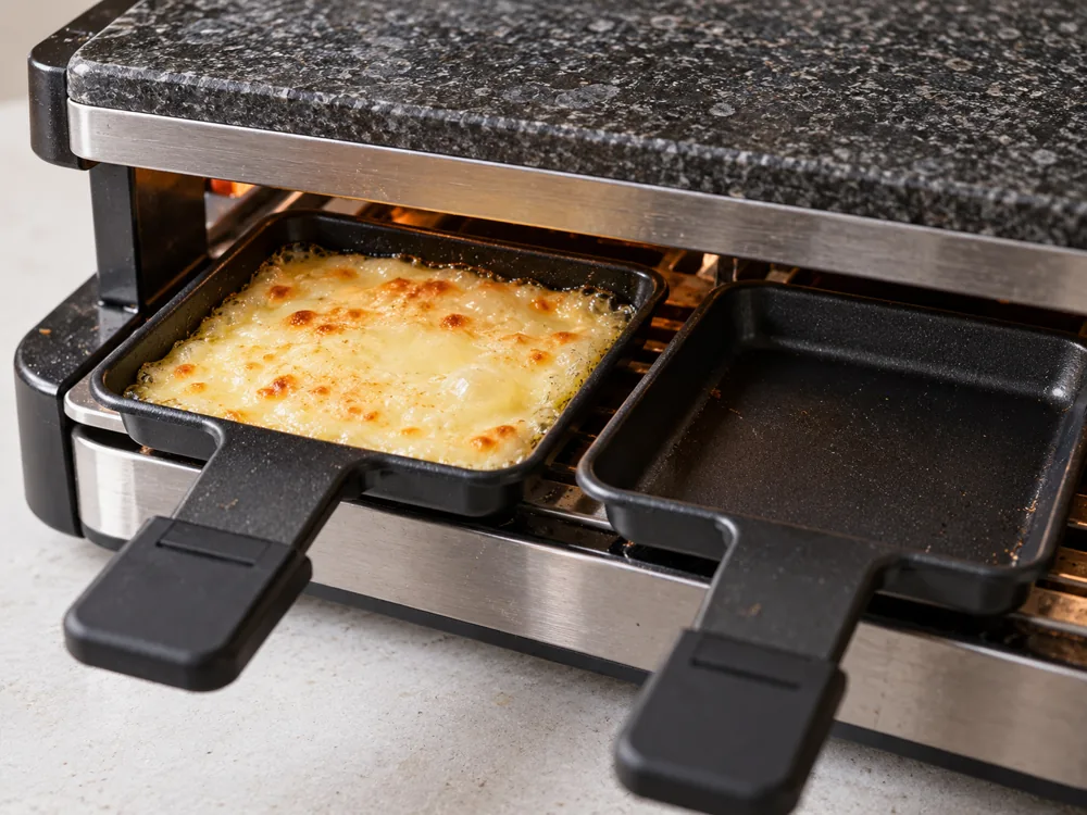 Slice of raclette melted in a small pan under a tabletop grill beside an empty pan - Raclette Cheese | KnowTheCheese