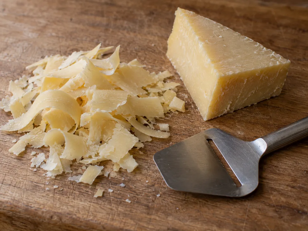 Pile of thin pale cheese shavings beside a rindless wedge and a flat cheese plane on a wooden board - Raclette Cheese | KnowTheCheese