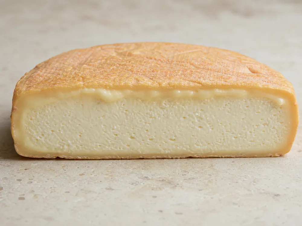 Reblochon cross-section with a thin washed rind, soft creamline, and ivory center - Reblochon | KnowTheCheese