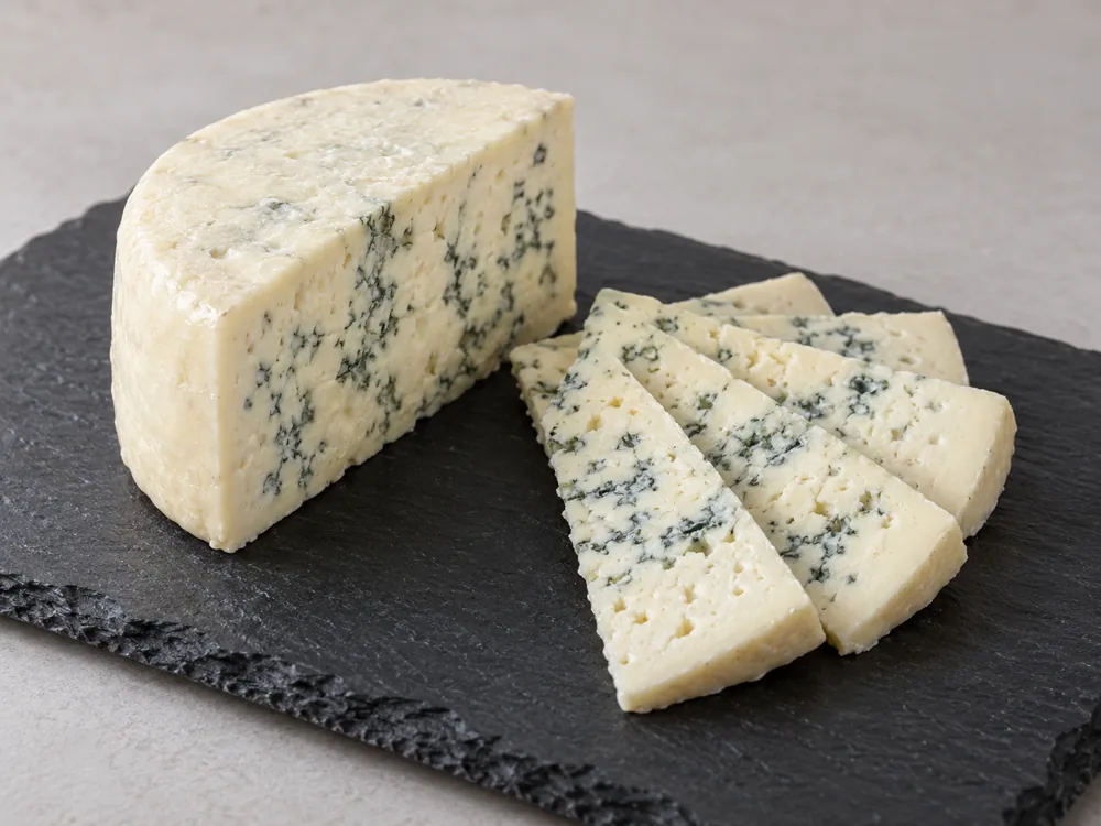 A large piece of blue-veined cheese beside thin fan-shaped slices on a slate board - Roquefort | KnowTheCheese